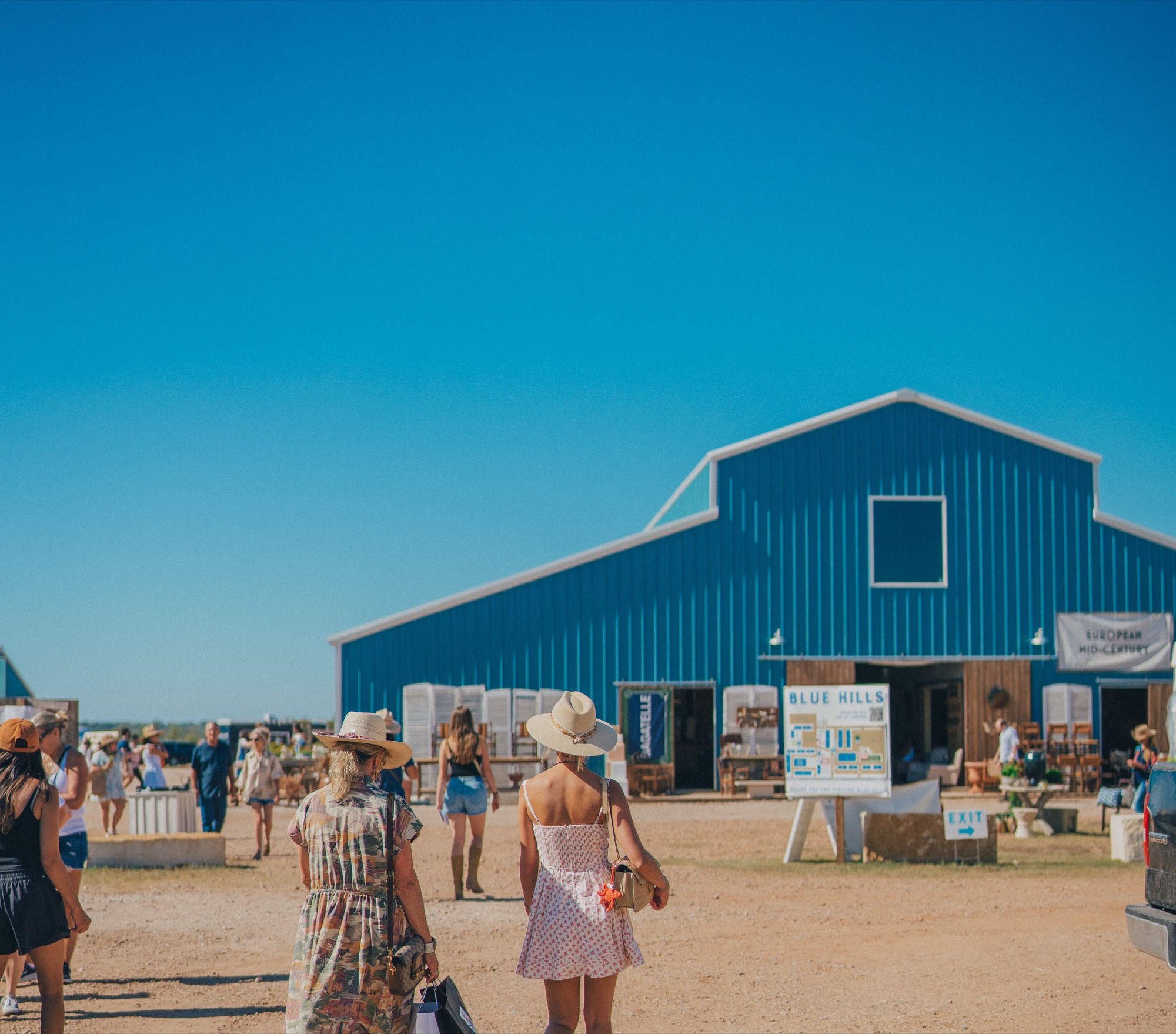 Two shoppers in sun hats walking toward the big blue Blue Hills barn under a clear Texas sky