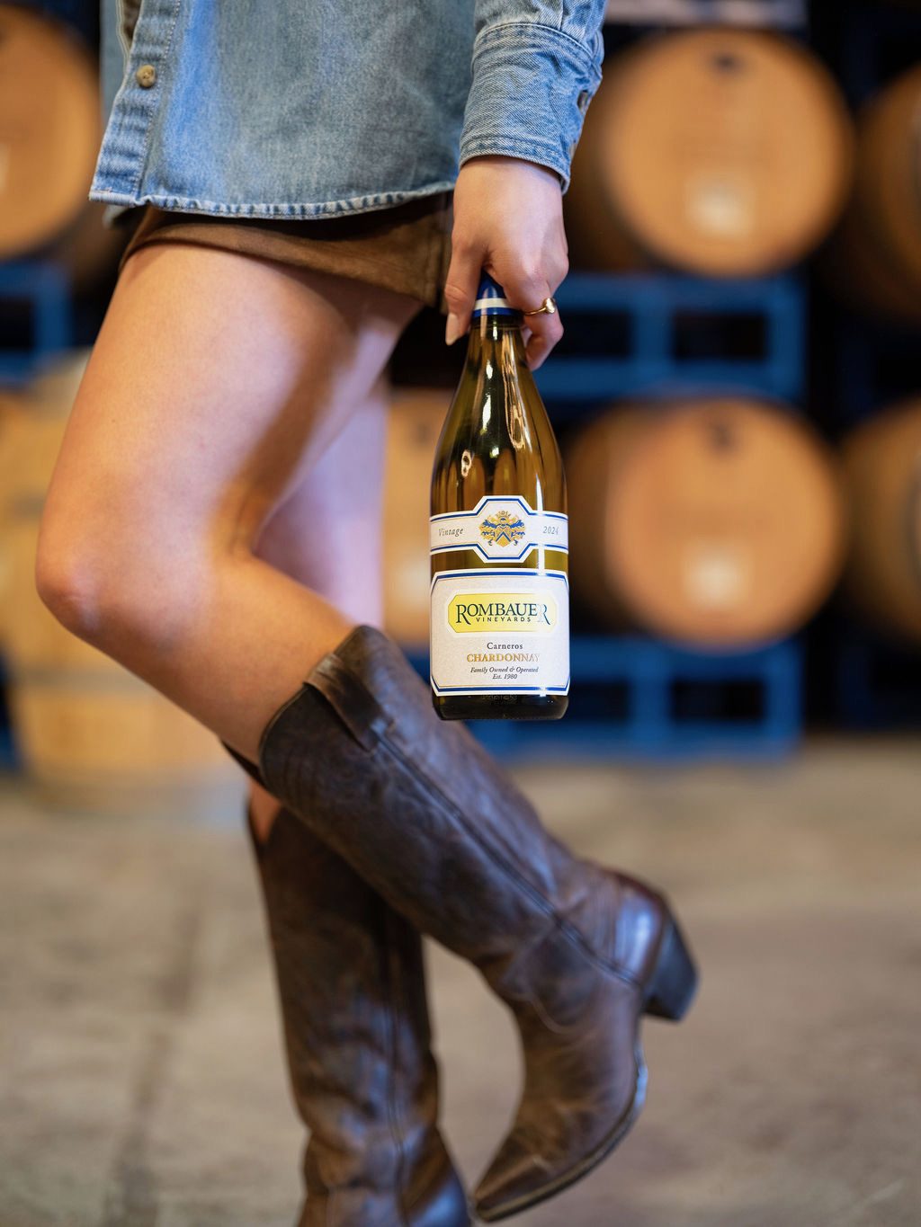 A woman in a denim shirt and tall cowboy boots carrying a bottle of Rombauer Carneros Chardonnay past stacked wine barrels