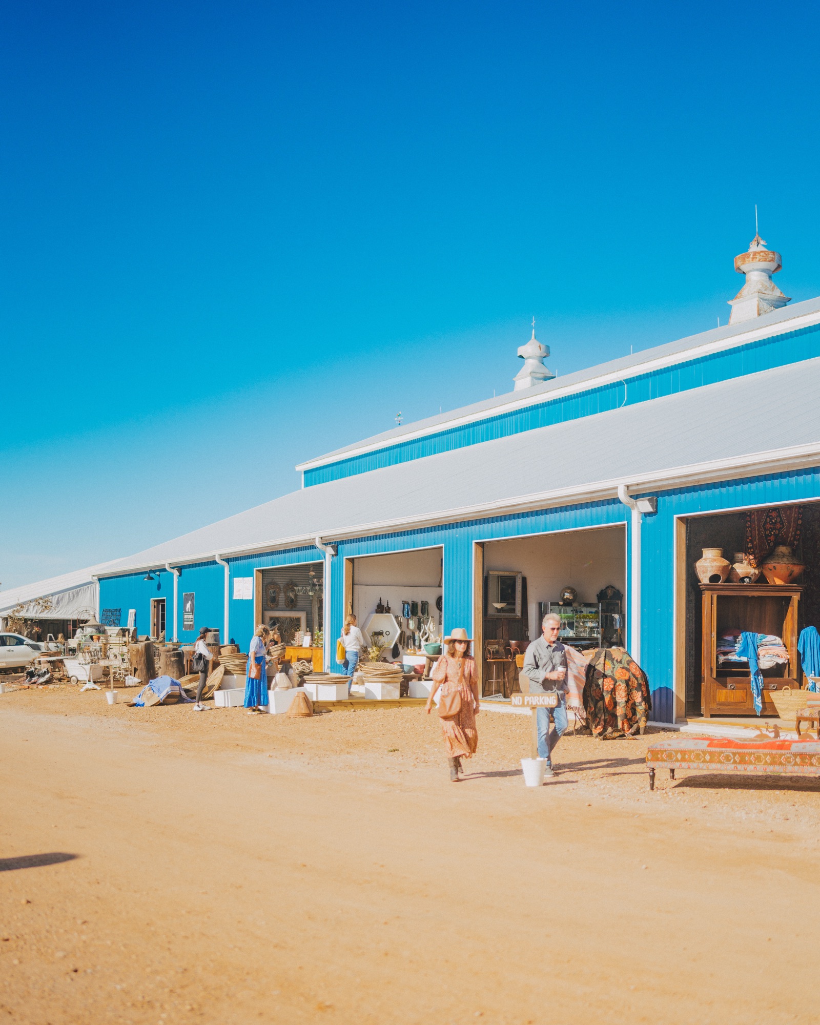 Shoppers browsing antiques in front of the bright-blue pole barns at Blue Hills on a sunny day